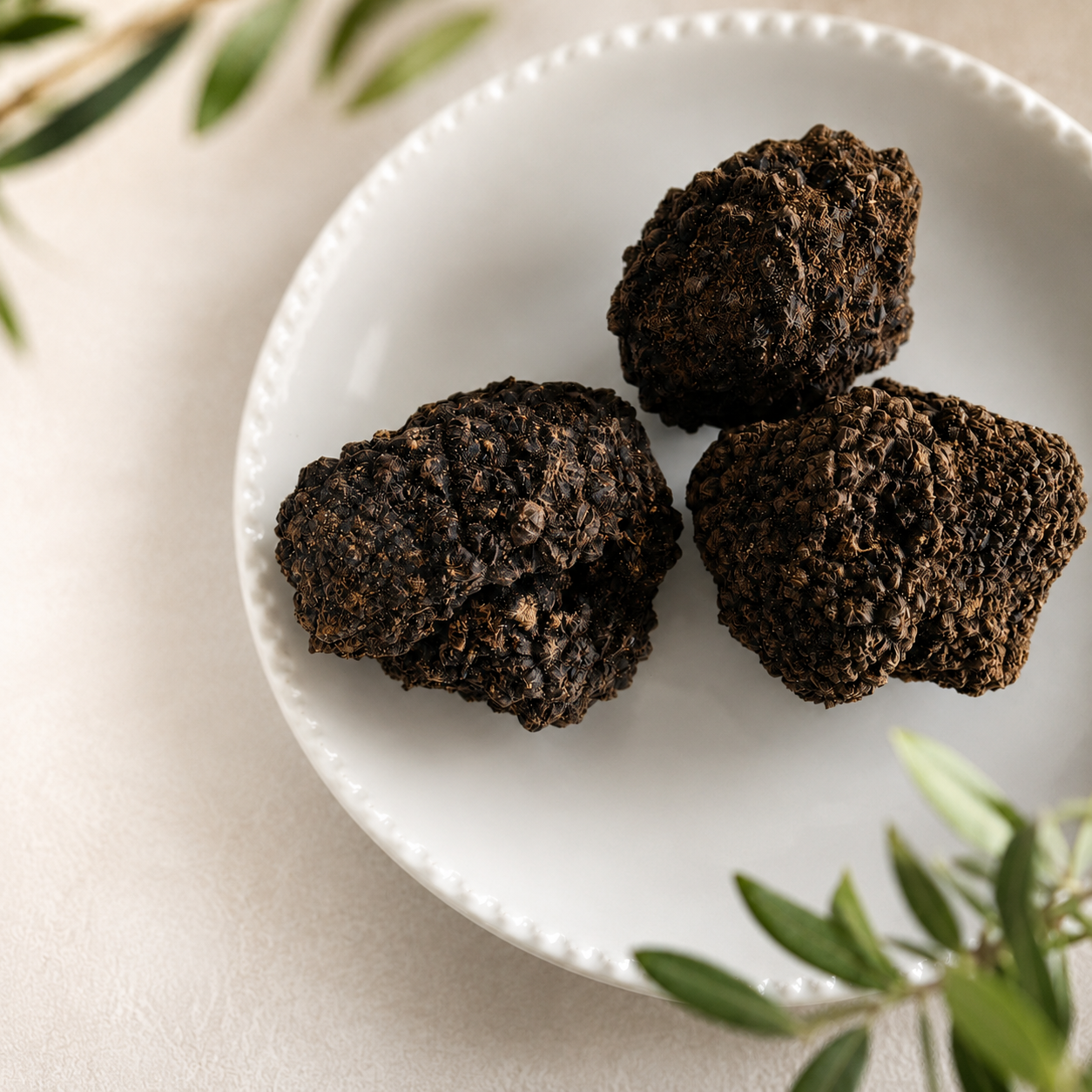 Tre tartufi neri ricoperti di granelli scuri su piatto bianco, con foglie verdi sullo sfondo.